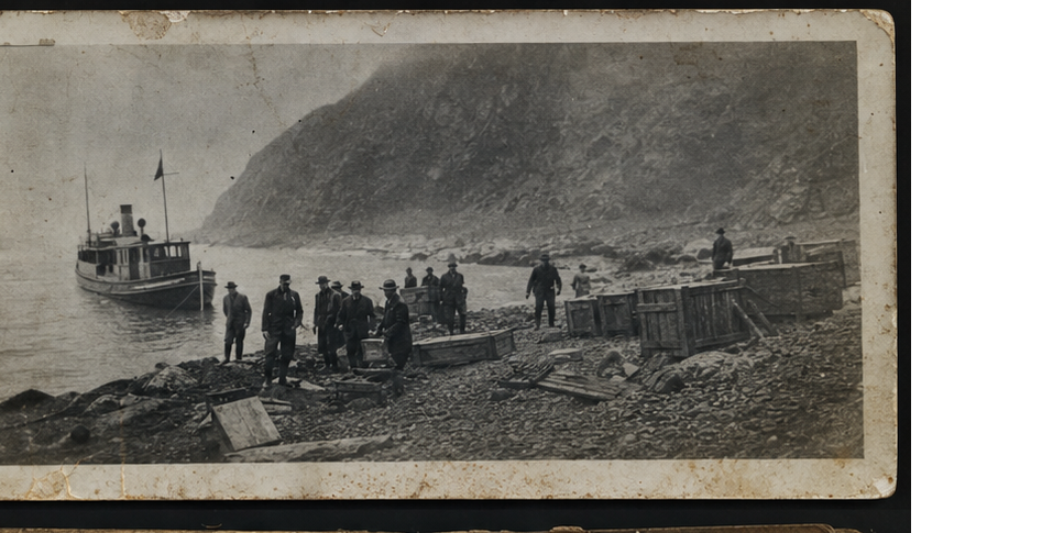 Poor-condition black and white photograph of a landing party, crates, and a small boat on a rocky island shore.
